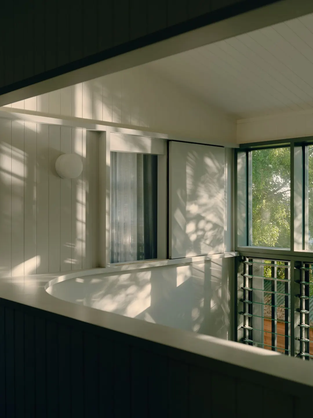 Curved white landing upstairs, with dappled light on VJ boards and glass louvres looking out to the trees