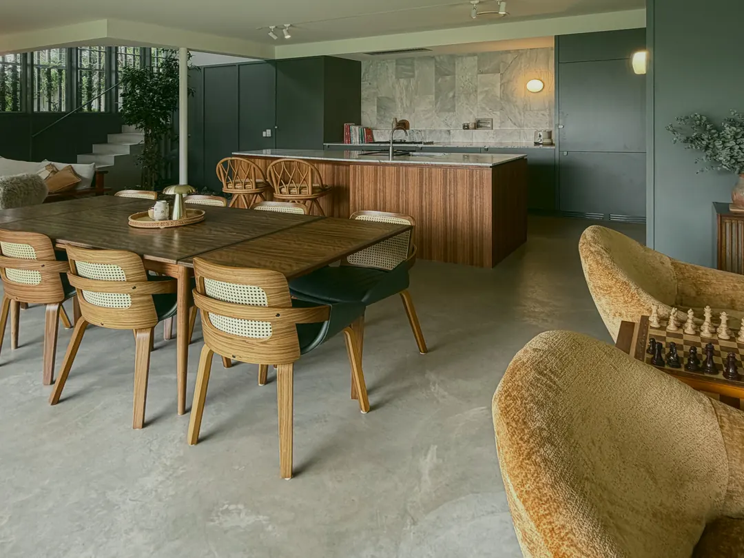 Cane-back dining chairs around a timber table, rattan stools at the spotted gum kitchen island, and a swivel armchair in the foreground