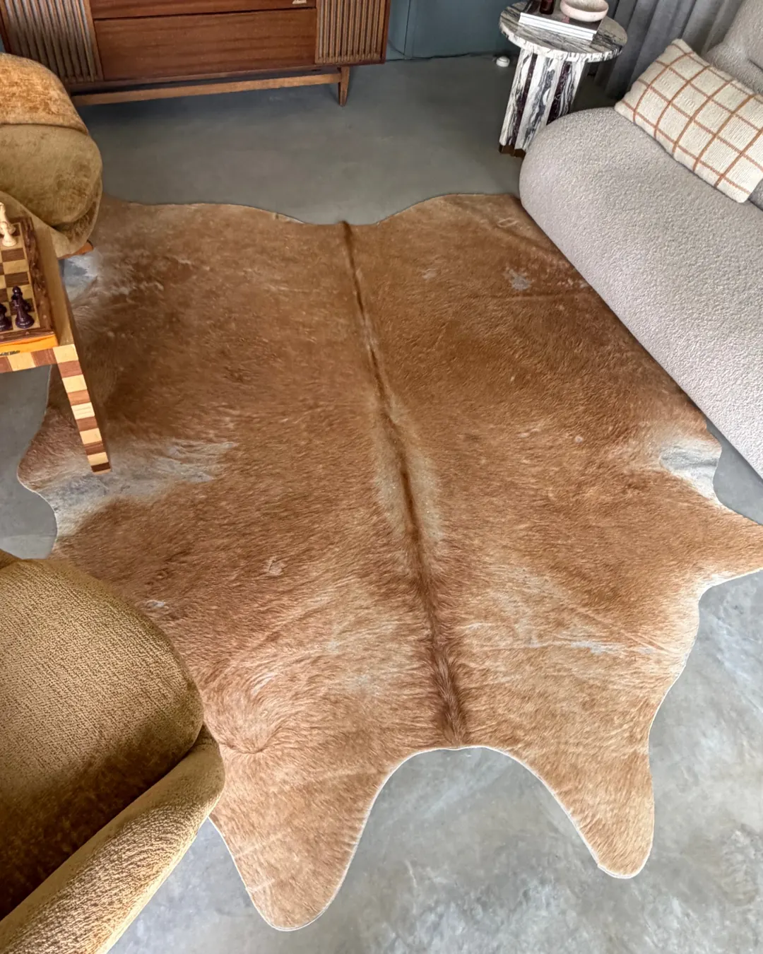 A natural tan cowhide rug on polished concrete, between swivel armchairs and a boucle sofa