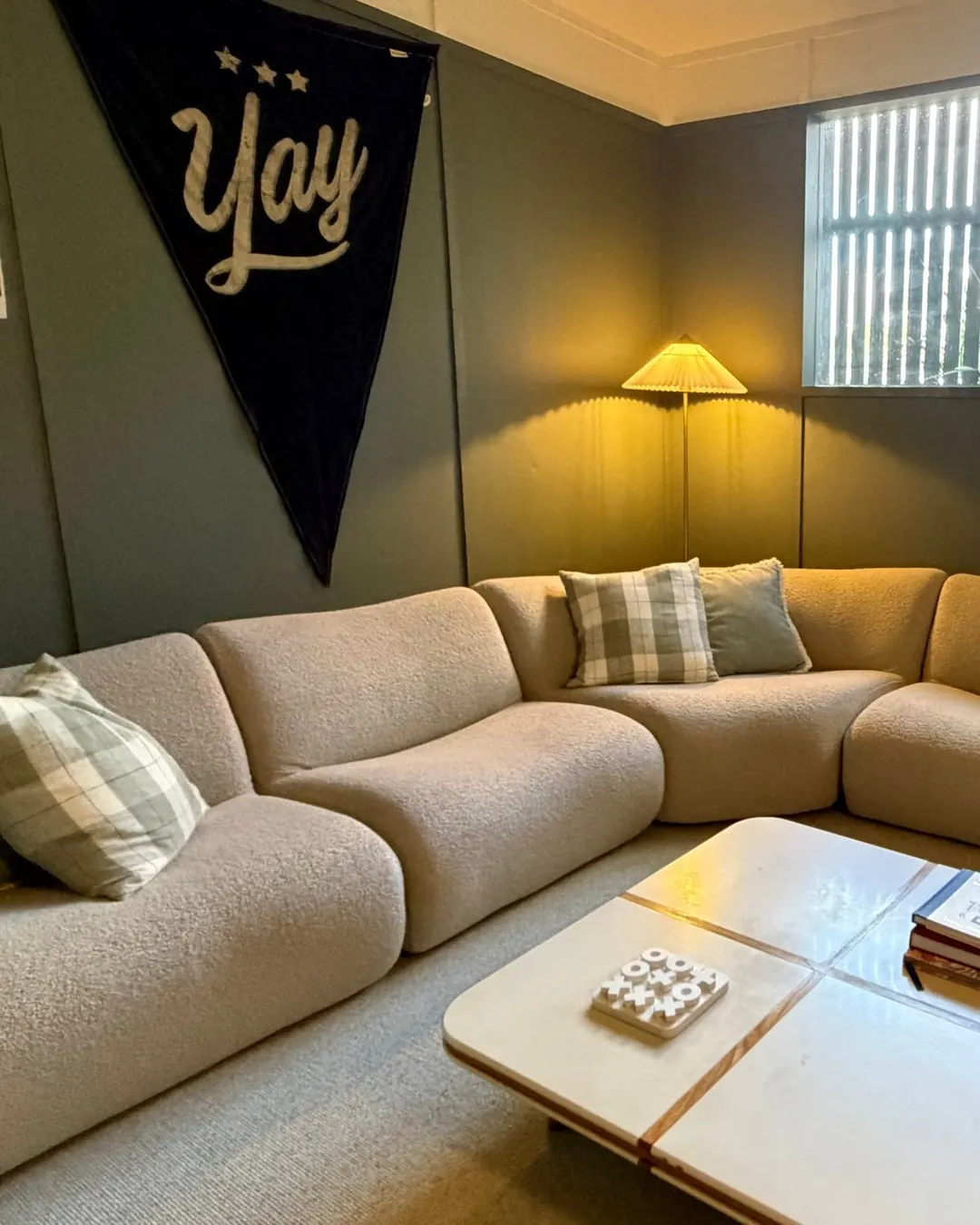 A modular King Living 1977 sofa in cream boucle wrapping the corner of the back room, with check cushions, a pleated floor lamp and a white tiled coffee table