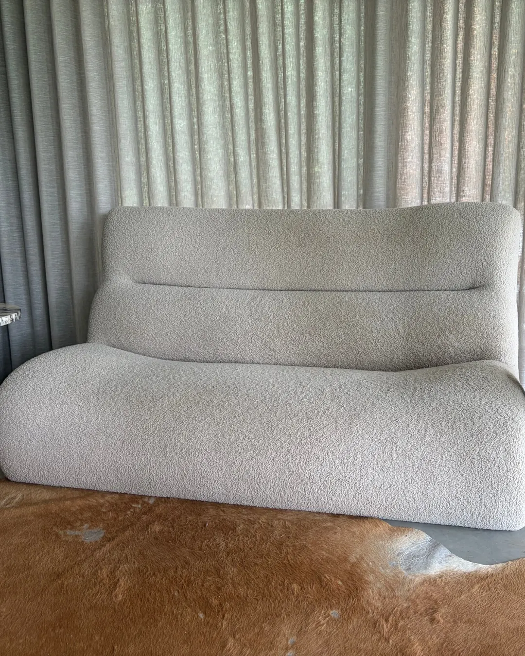 A high-back boucle sofa in oatmeal in front of linen curtains, with a cowhide rug at its feet