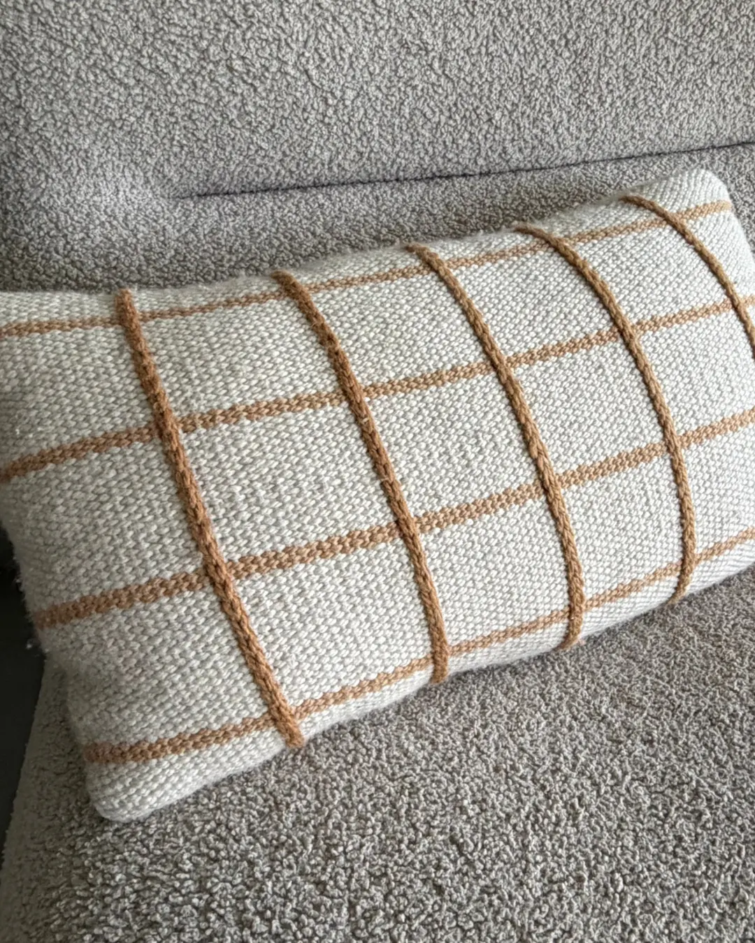 A rectangular woven cushion in cream with a tan grid, on a boucle sofa