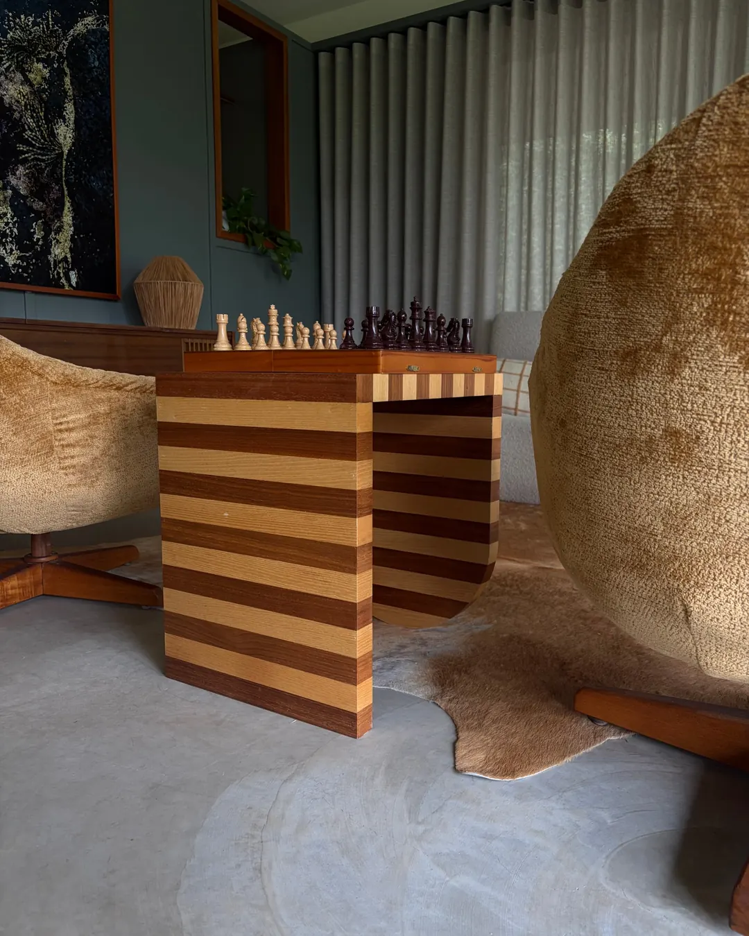 A side table in striped ash and walnut with a chess set on top, between two swivel armchairs on a cowhide rug