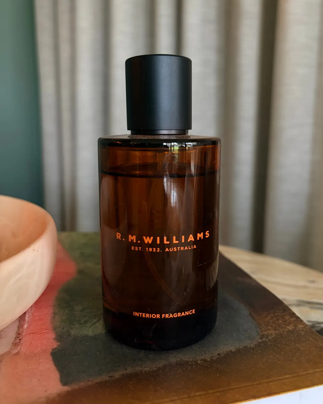 An amber glass bottle of R.M.Williams interior fragrance on a marble side table, with a linen curtain behind