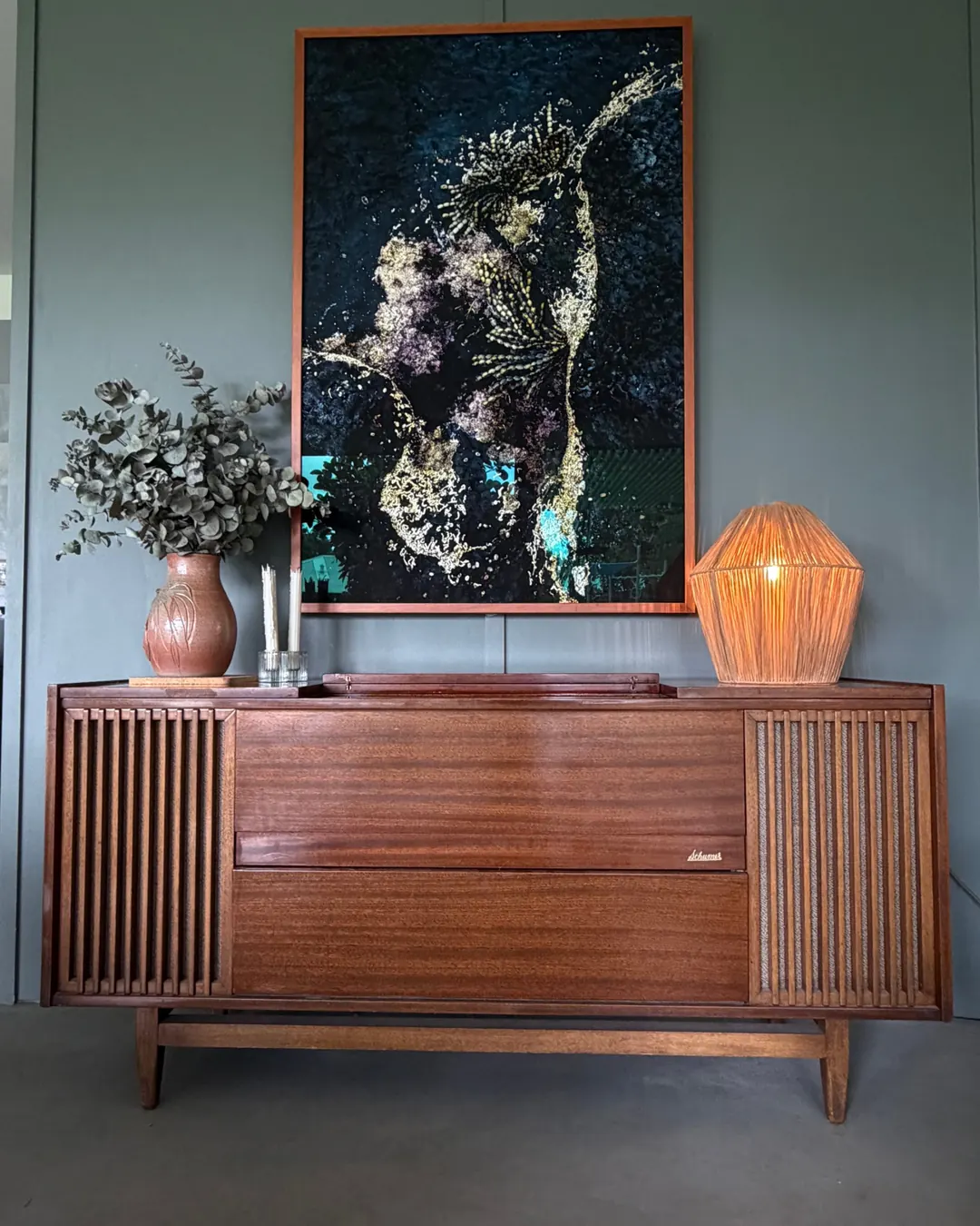 A Schumer radiogram in timber with slatted speaker ends, a raffia lamp and a vase of eucalyptus on top, and a framed artwork above