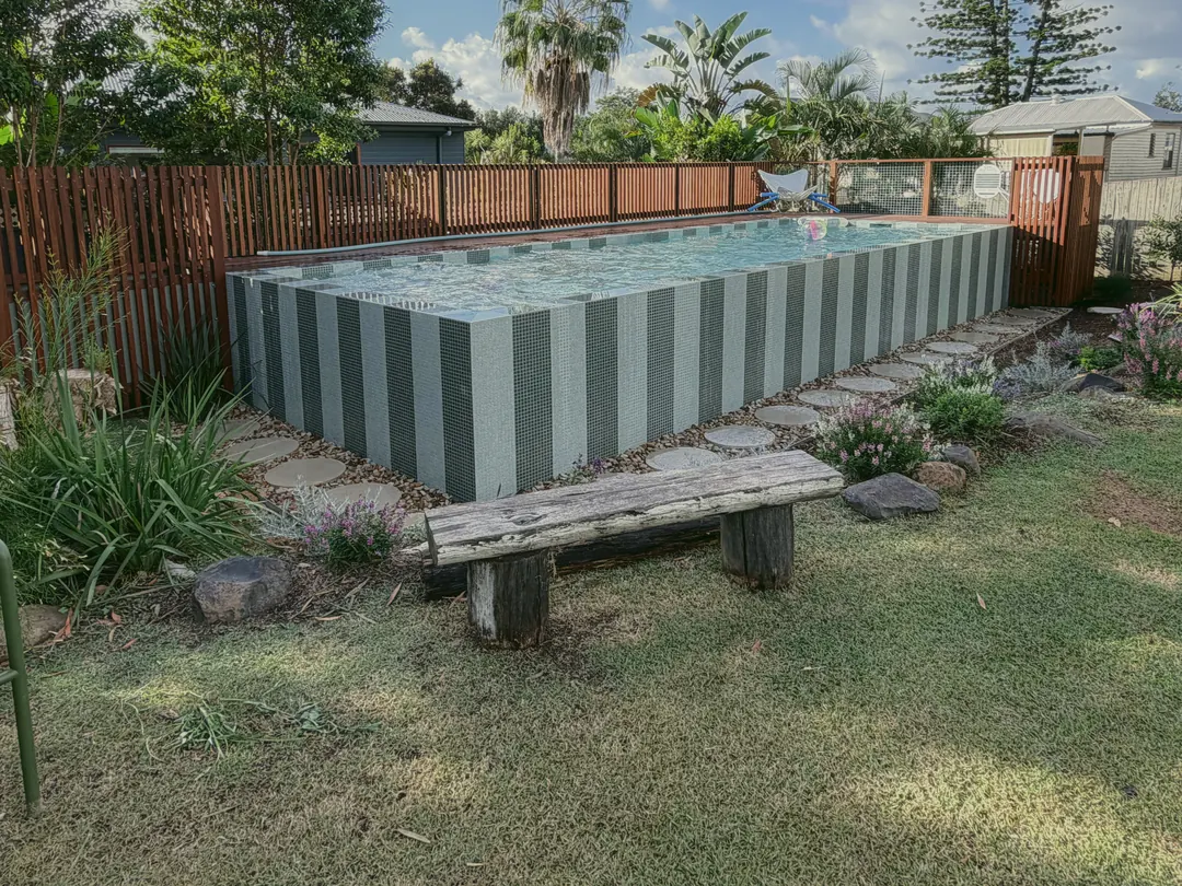 A striped green-tiled plunge pool behind a timber batten fence, stepping stones and flowering natives around it, an old timber bench on the lawn