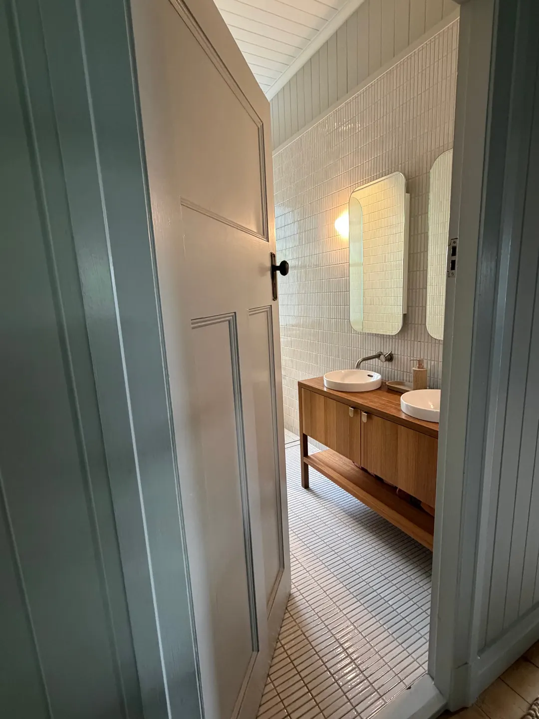 Looking through a panelled door into a bright white bathroom in Japanese finger tiles, with a spotted gum vanity, two round basins and rounded mirrors